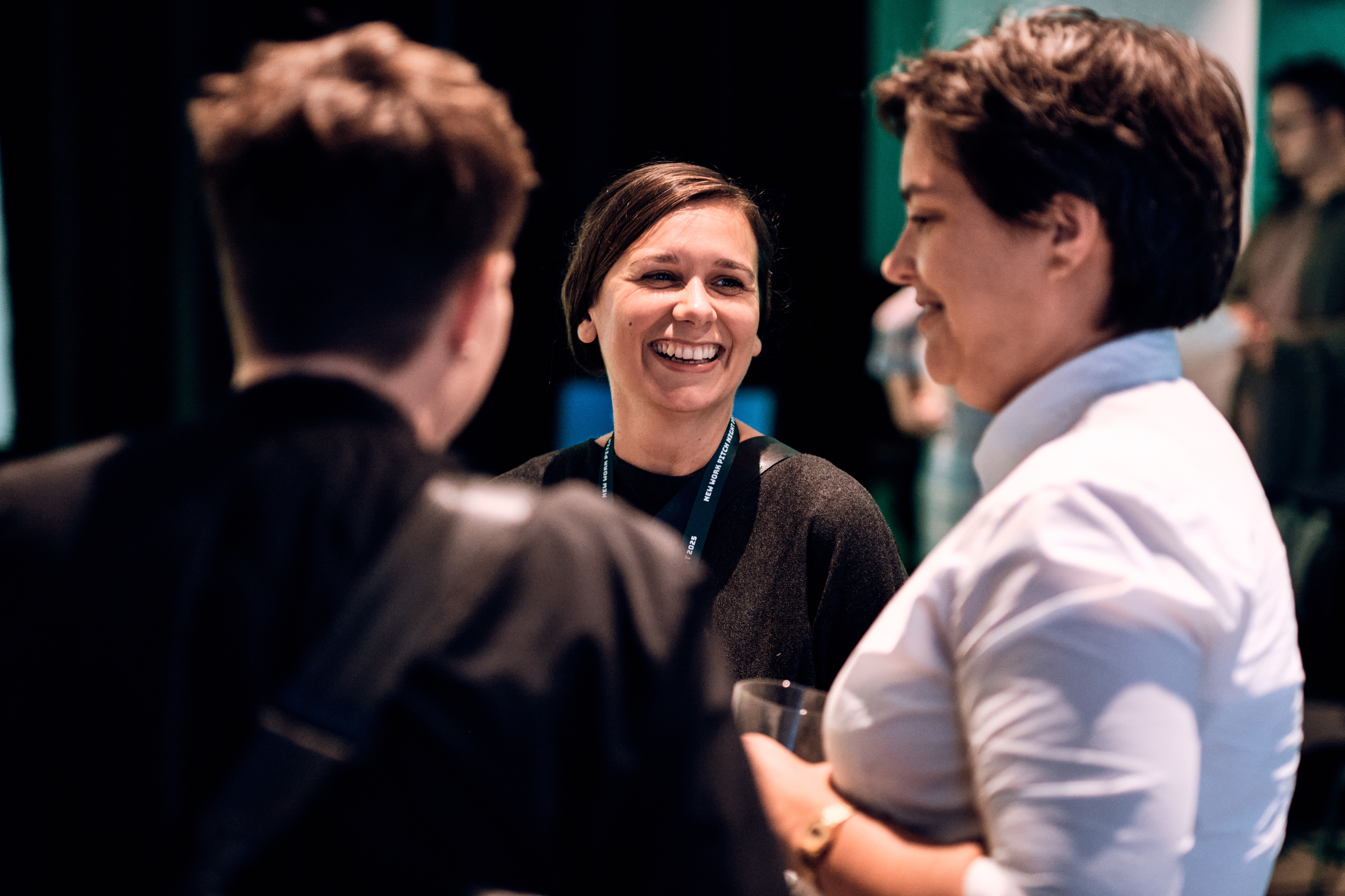 Drei Personen stehen bei einer Indoor-Veranstaltung zusammen und unterhalten sich; die Person in der Mitte lächelt, während die anderen ihr zugewandt sind.